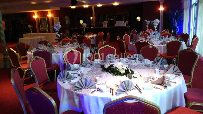 Salle de réception avec tables rondes dressées, nappes blanches, serviettes pliées et chaises rouges.