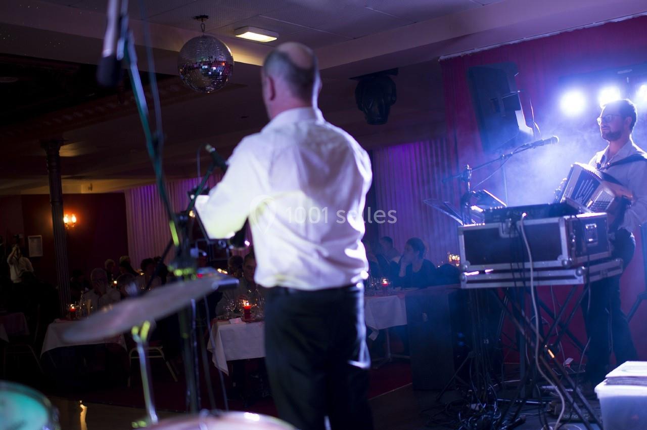 Un musicien joue de l'accordéon sur scène dans une salle éclairée, devant un public assis à des tables.