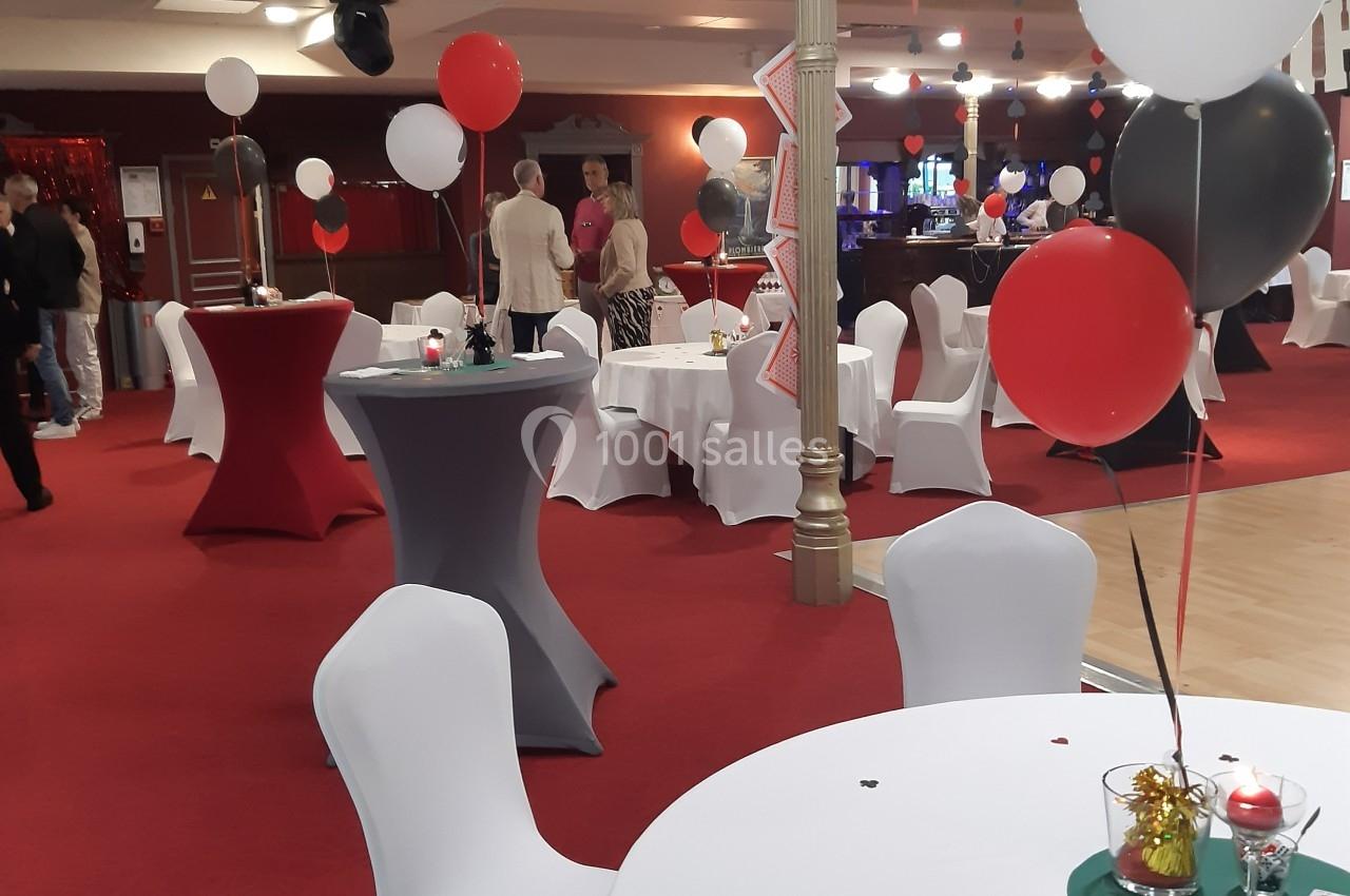 Salle décorée avec des ballons rouges, noirs et blancs, tables rondes couvertes de nappes blanches, ambiance festive.