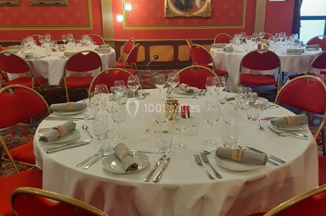 Salle élégante avec tables rondes dressées, nappes blanches, serviettes pliées et chaises rouges, décorée de tableaux.
