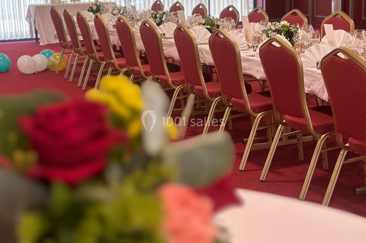 Salle de réception avec tables dressées, nappes blanches, chaises rouges et décorations florales au premier plan.