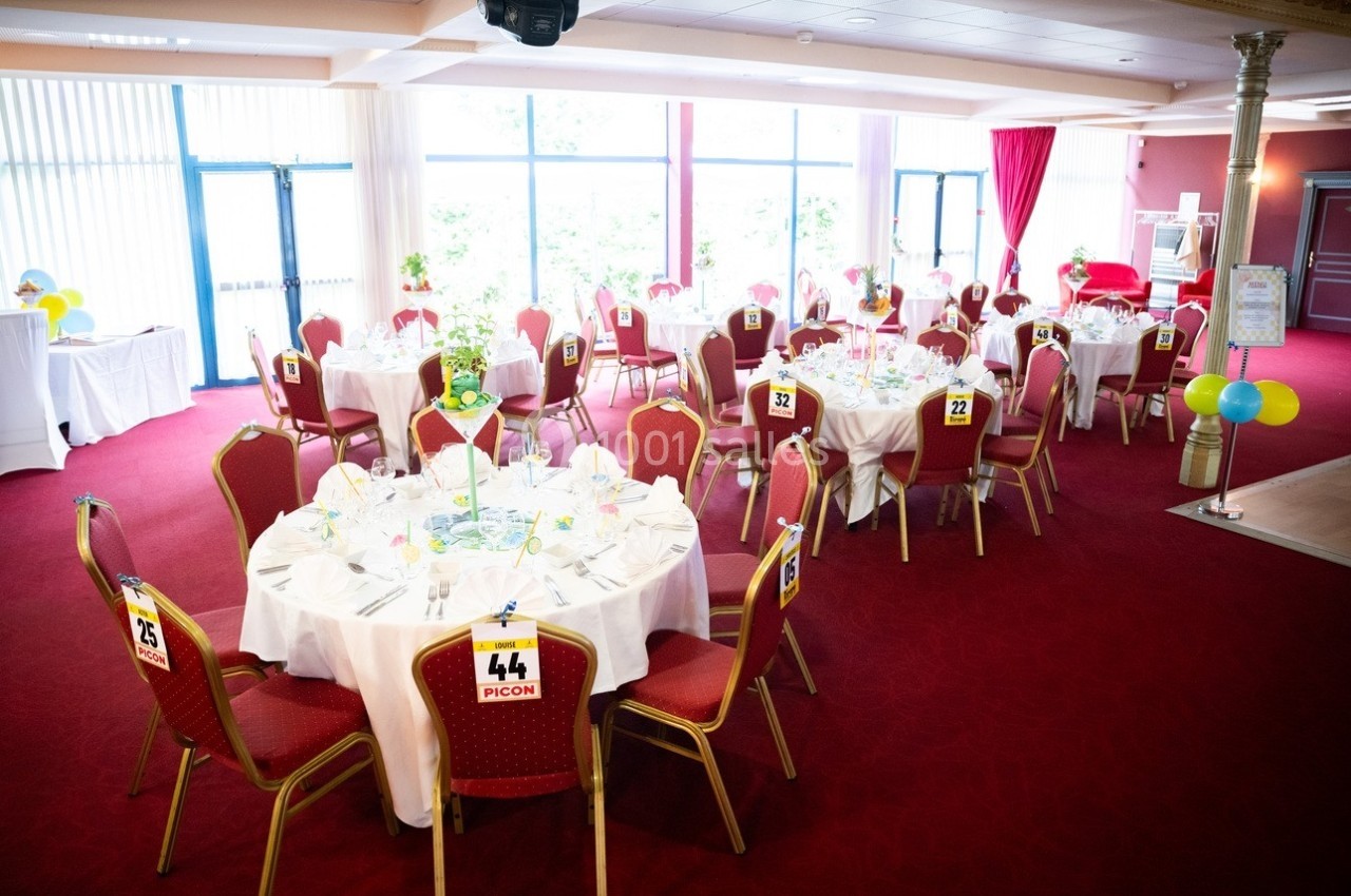 Salle de réception avec tables rondes dressées, nappes blanches, chaises rouges et numéros de table visibles.
