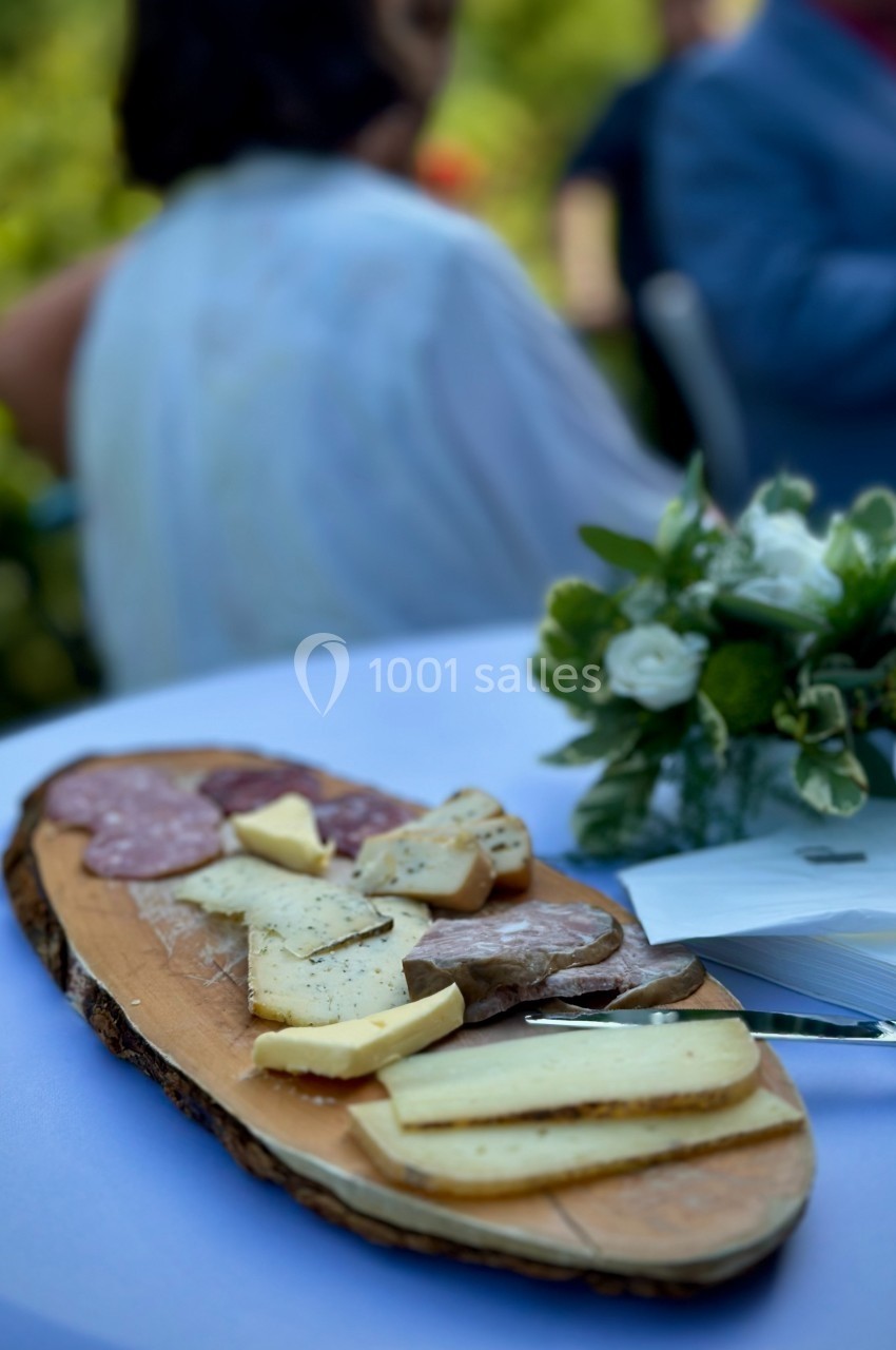 Plateau de charcuterie et de fromages variés sur une planche en bois, posé sur une table blanche avec un bouquet de fleurs.