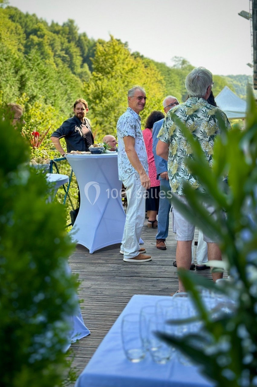 Des personnes discutent debout autour de tables hautes sur une terrasse en bois entourée de verdure.