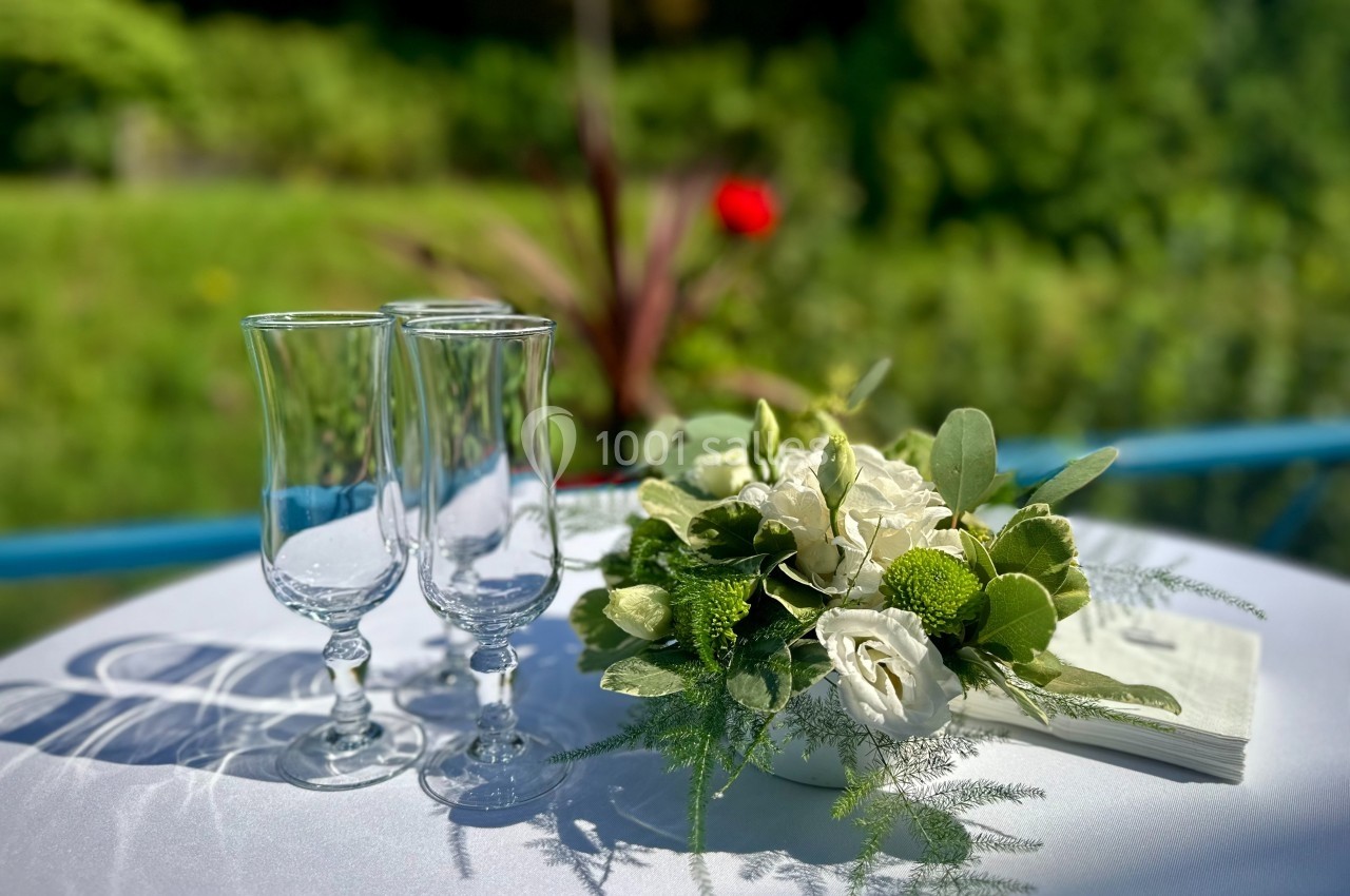 Deux verres à pied et un bouquet de fleurs blanches et vertes sur une table ronde en extérieur.