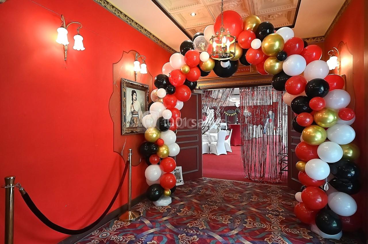Entrée décorée avec une arche de ballons rouges, noirs, blancs et dorés, menant à une salle ornée de rideaux et tables.