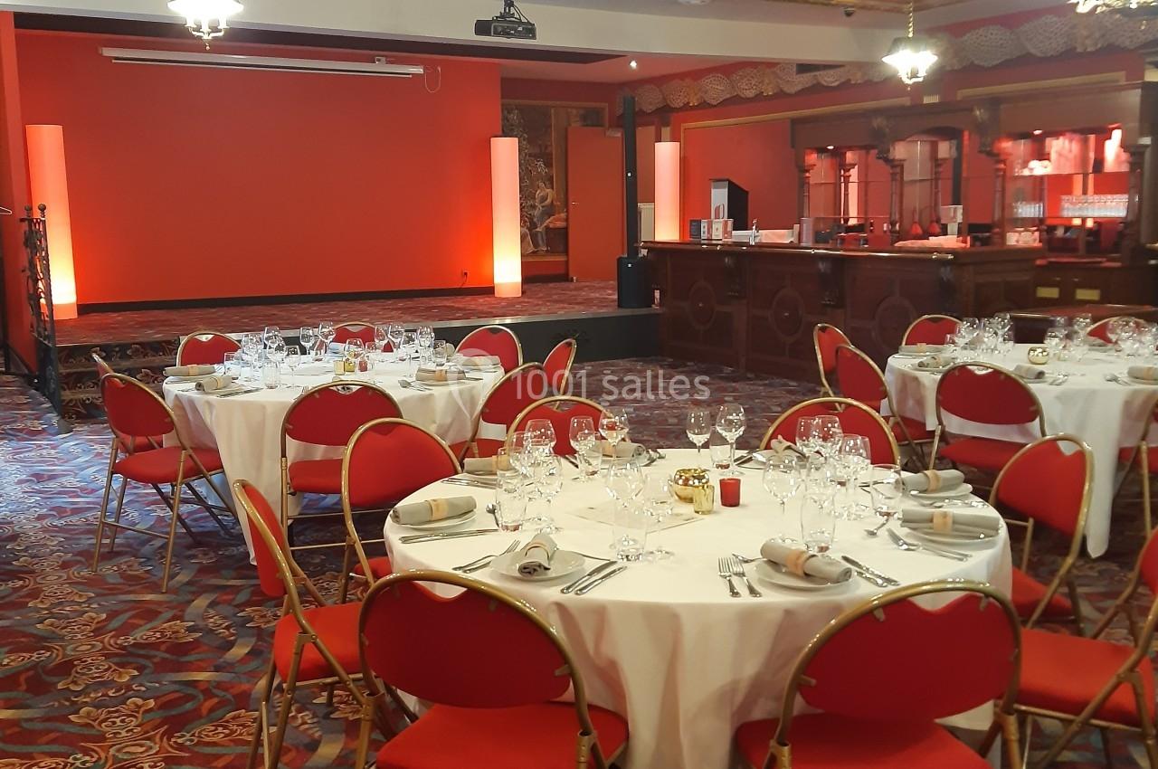 Salle de réception avec tables rondes dressées, chaises rouges et éclairage chaleureux près d'un bar en bois.