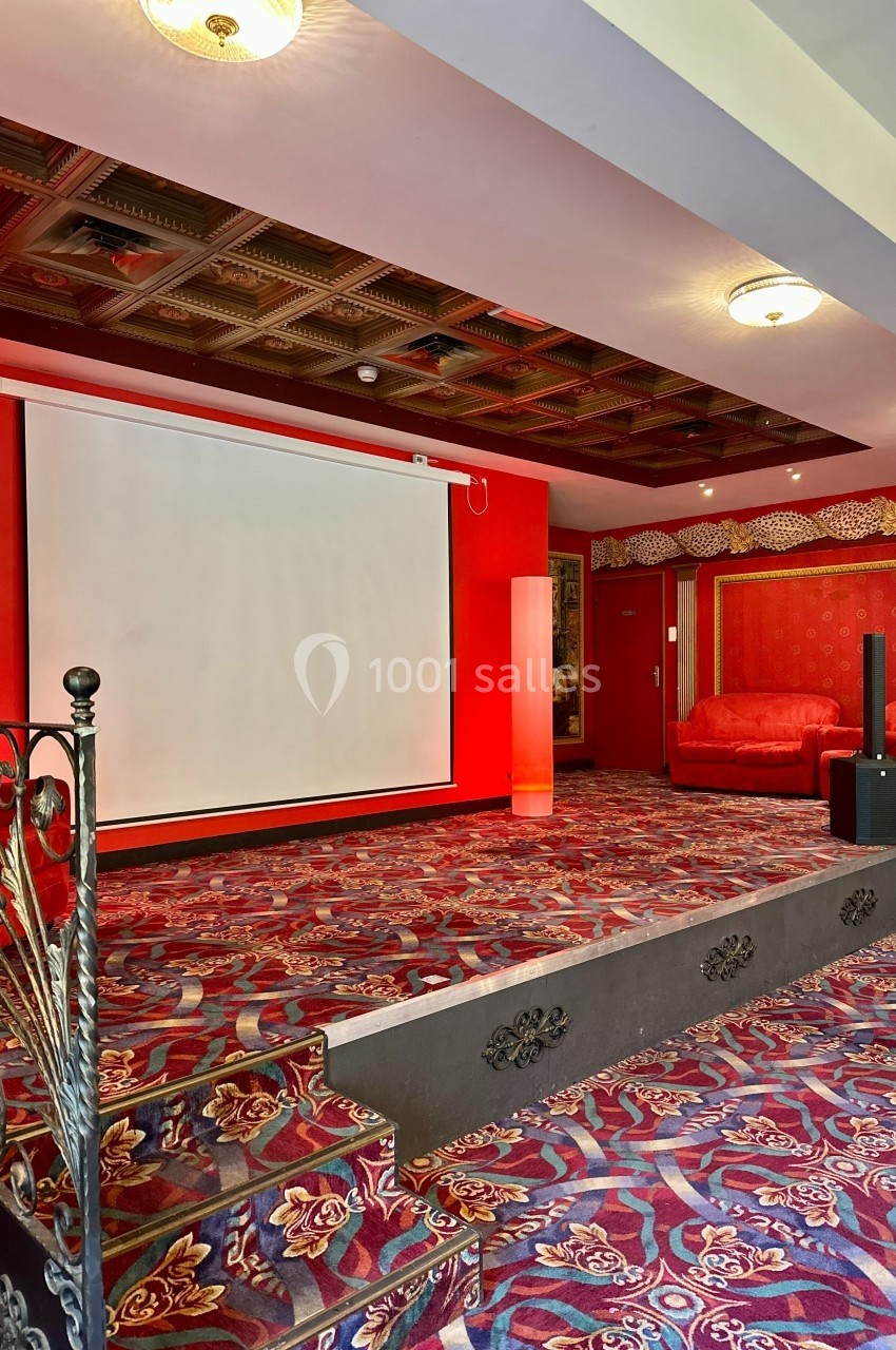 Salle avec écran de projection, tapis coloré, murs rouges, fauteuils et éclairage au plafond.