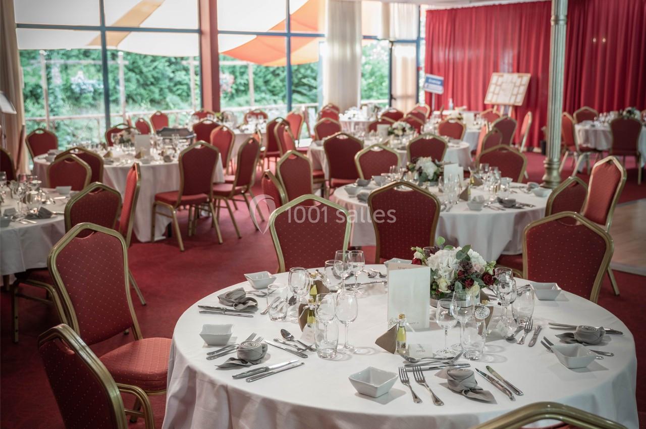 Salle de réception aménagée avec des tables rondes dressées, nappes blanches, chaises rouges et décorations florales.