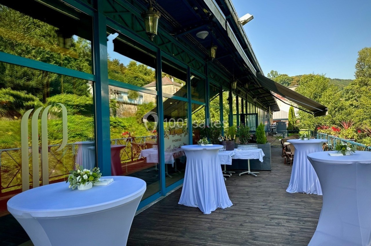 Terrasse en bois avec tables hautes drapées de blanc, entourée de verdure et bordée par une façade vitrée.