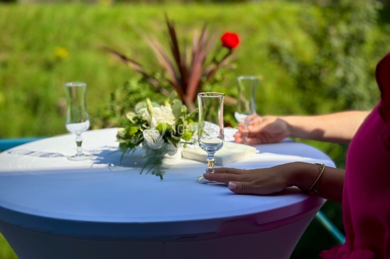 Deux mains posées sur une table haute blanche, décorée de fleurs, avec des verres vides en extérieur verdoyant.