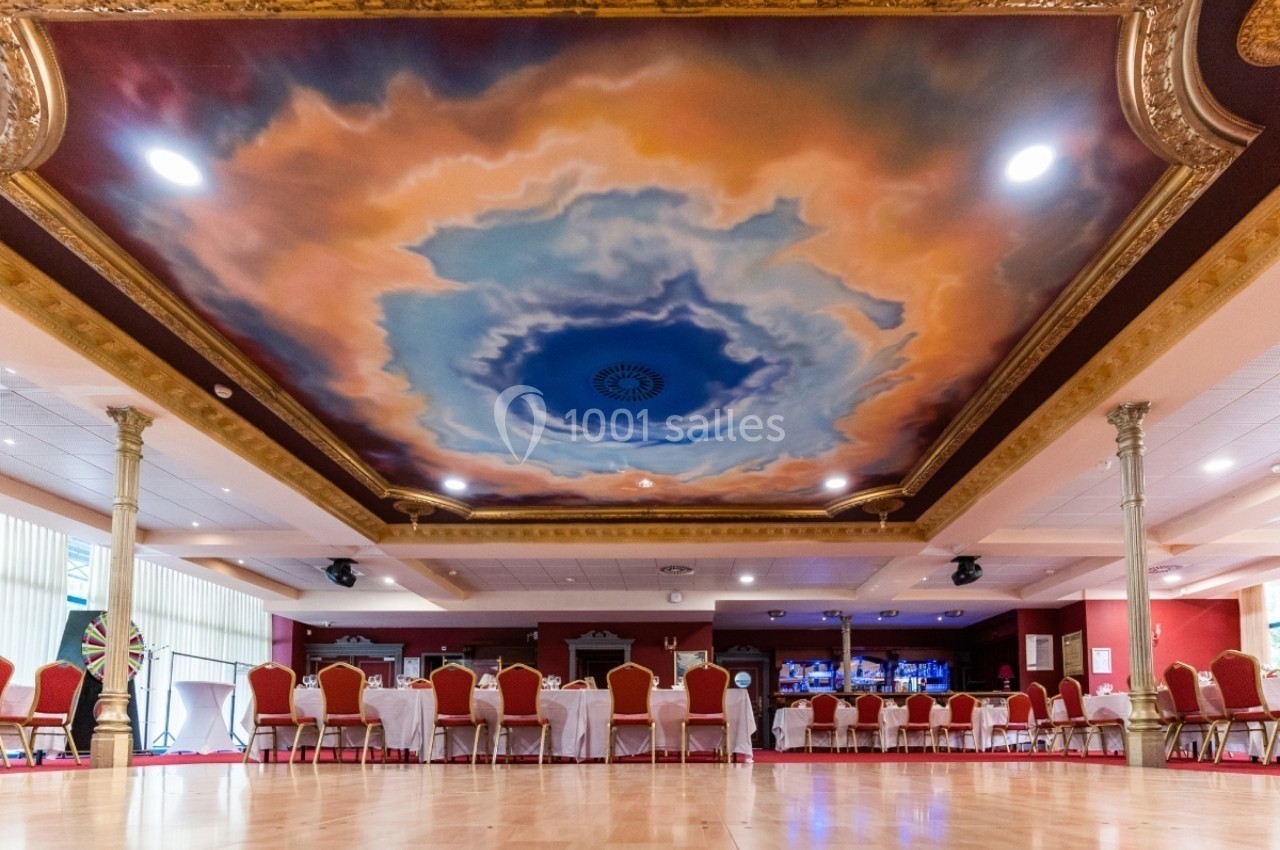Location salle Plombières-les-Bains (Vosges) - Casino de Plombières #2 Plafond peint avec un ciel nuageux entouré de moulures dorées dans une grande salle avec des chaises et tables alignées.