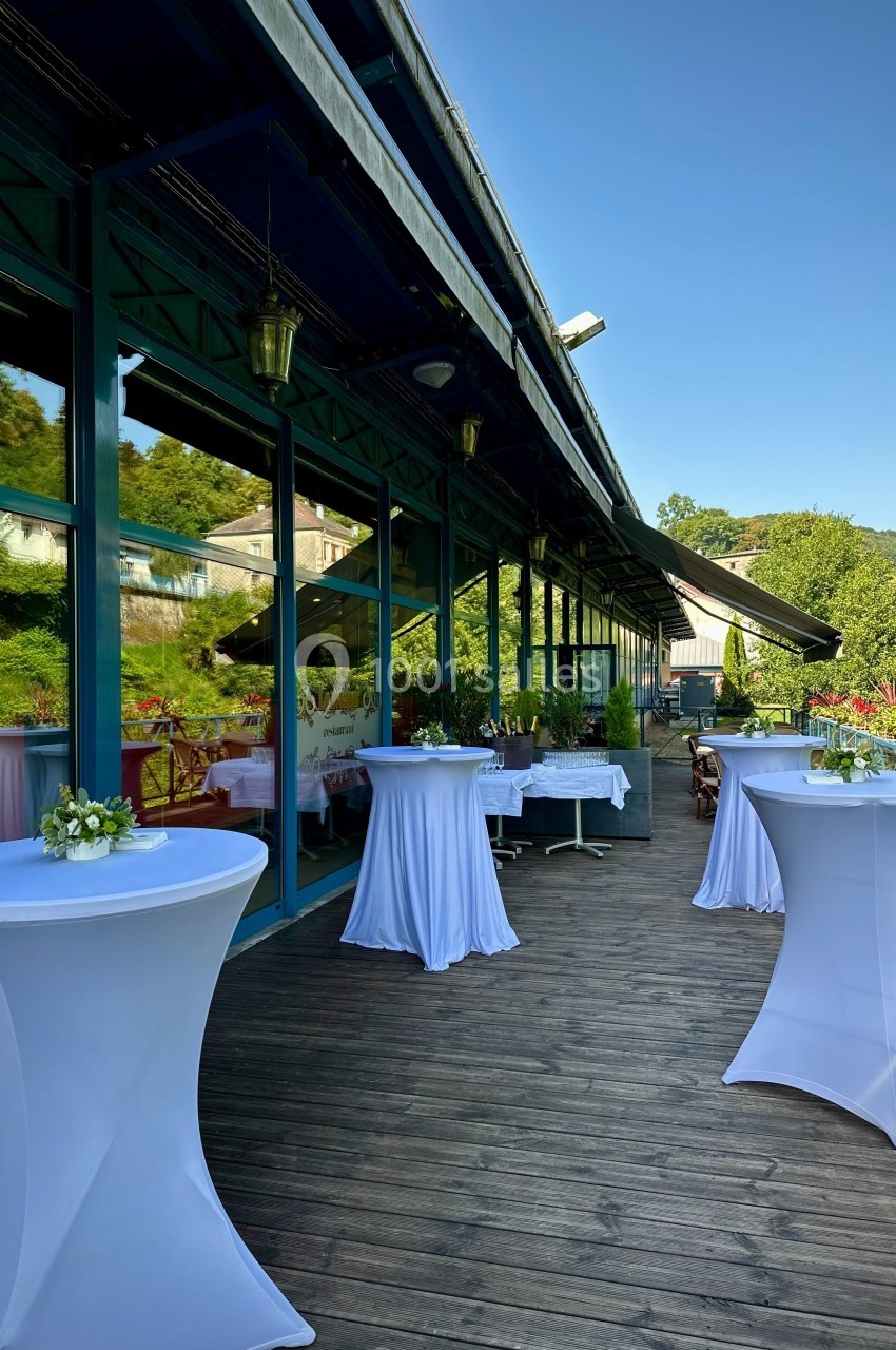 Location salle Plombières-les-Bains (Vosges) - Casino de Plombières #17 Terrasse en bois avec tables hautes drapées de blanc, entourée de verdure et jouxtant un bâtiment vitré.