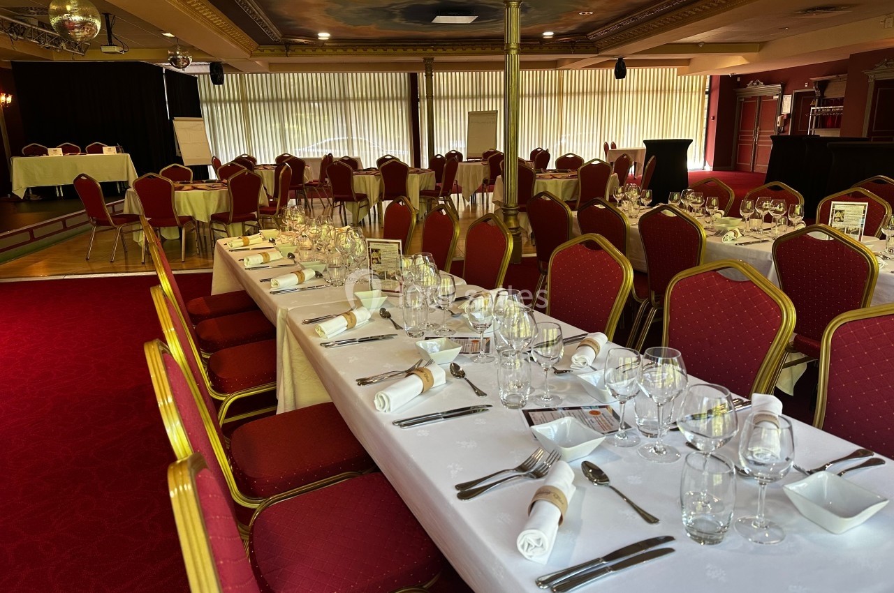Salle de réception avec tables dressées, nappes blanches, chaises rouges et lumière naturelle filtrée par des stores.
