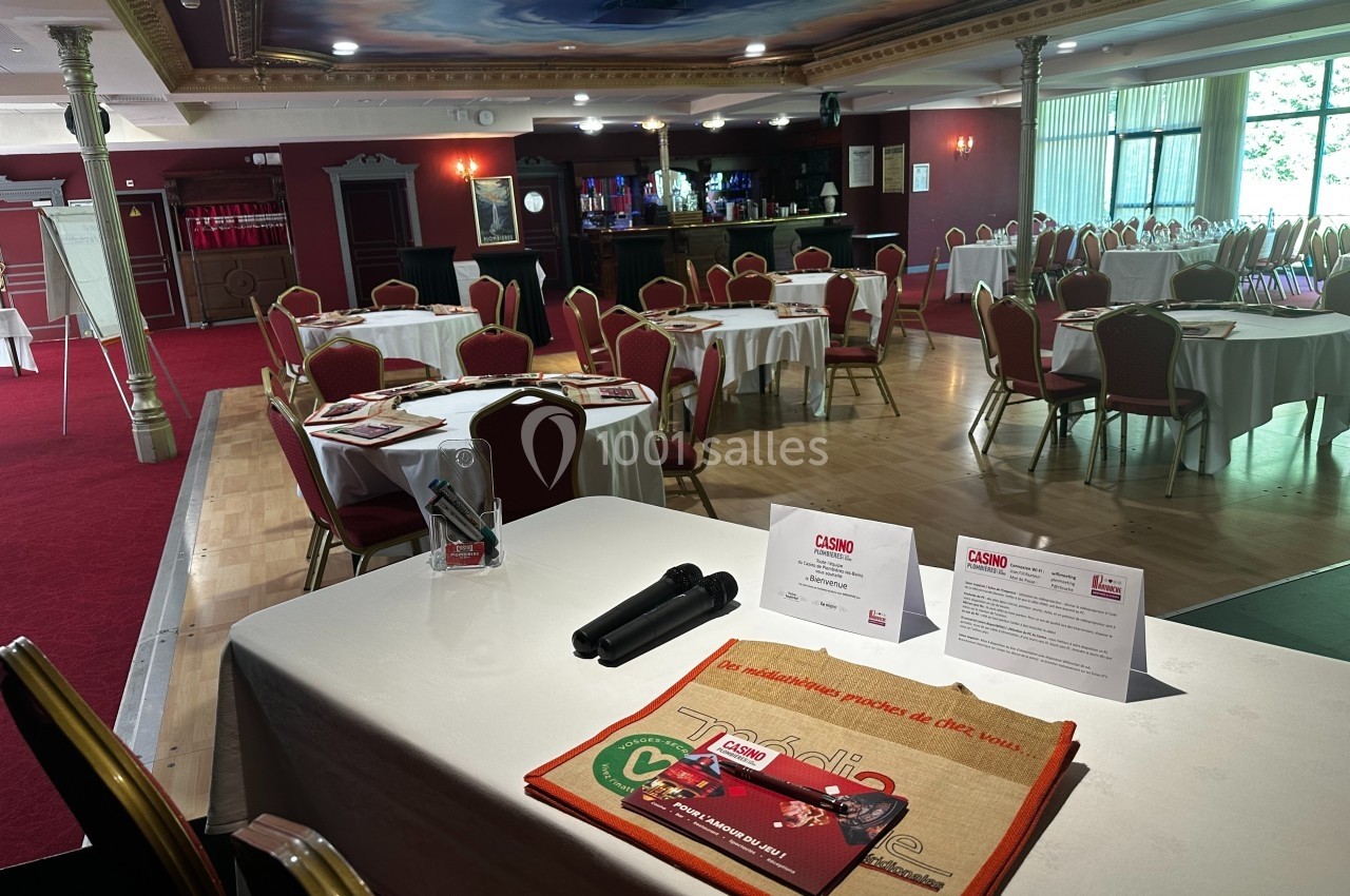Salle de conférence avec tables rondes dressées, documents promotionnels et micros sur une table au premier plan.