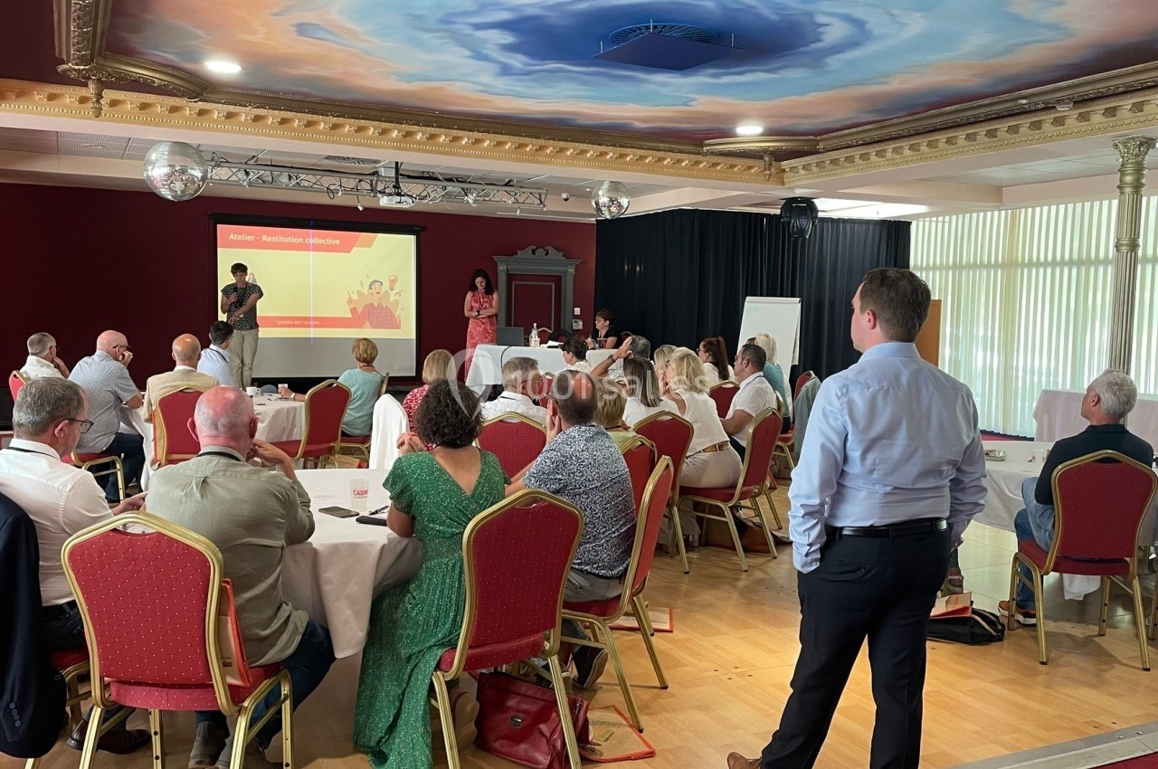 Groupe de personnes assises dans une salle de conférence, écoutant une présentation projetée sur un écran.