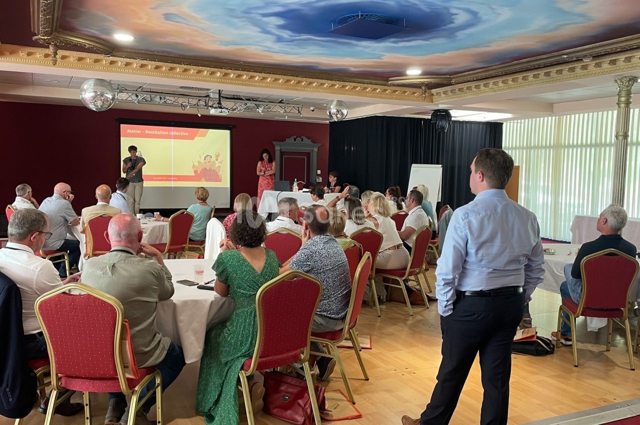 Groupe de personnes assises dans une salle de conférence, écoutant une présentation projetée sur un écran.