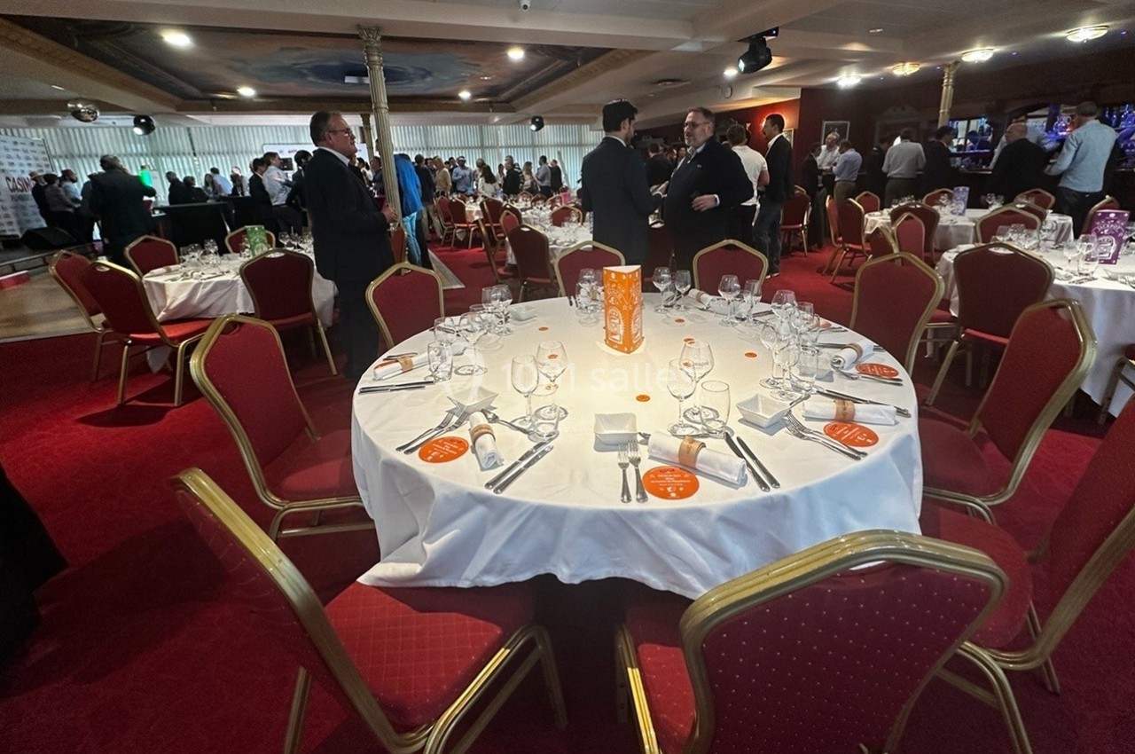 Salle de réception avec tables rondes dressées, nappes blanches, chaises rouges et invités debout en arrière-plan.