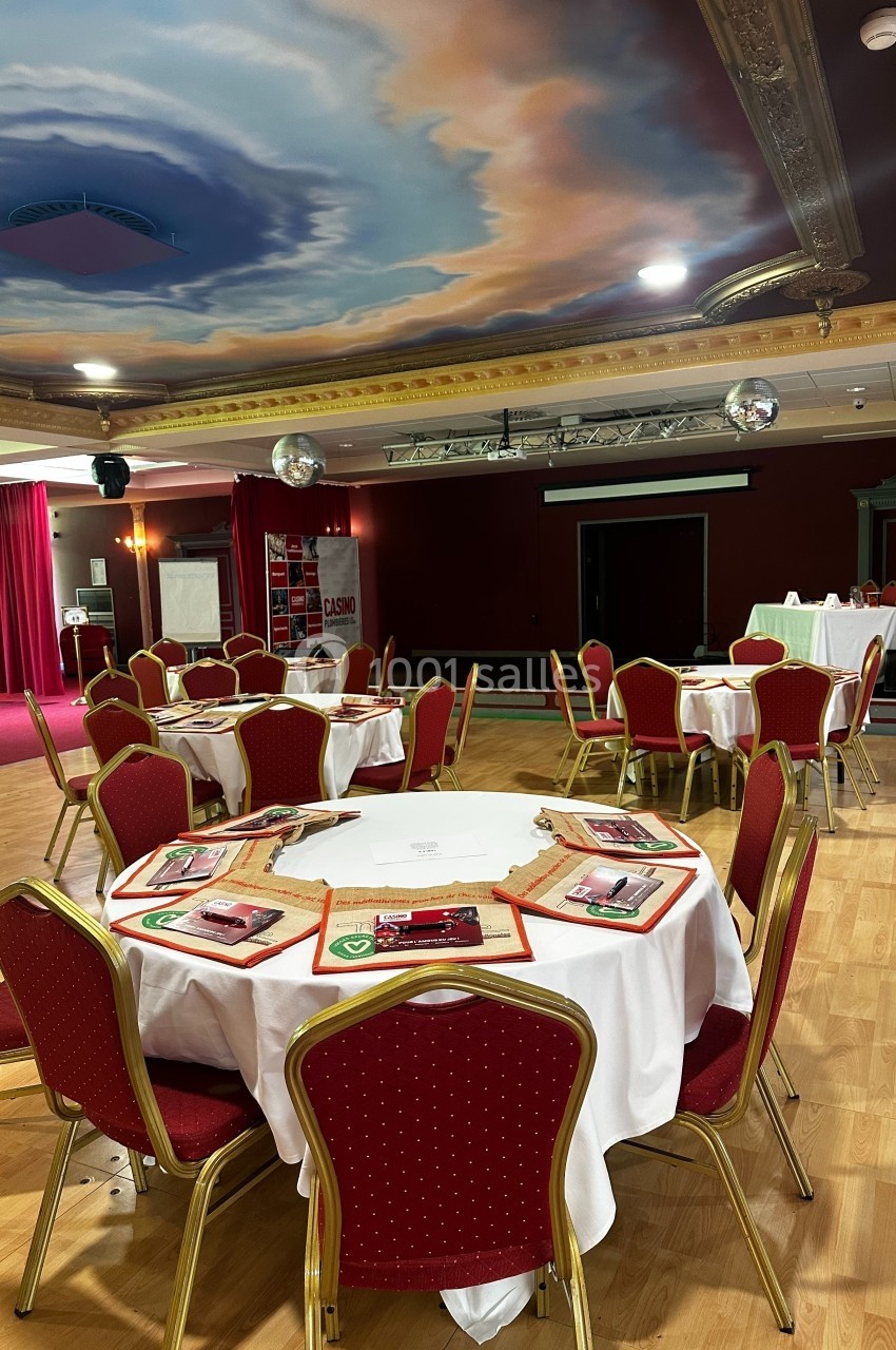 Salle de réception avec tables rondes dressées, nappes blanches, chaises rouges et plafond peint en ciel nuageux.