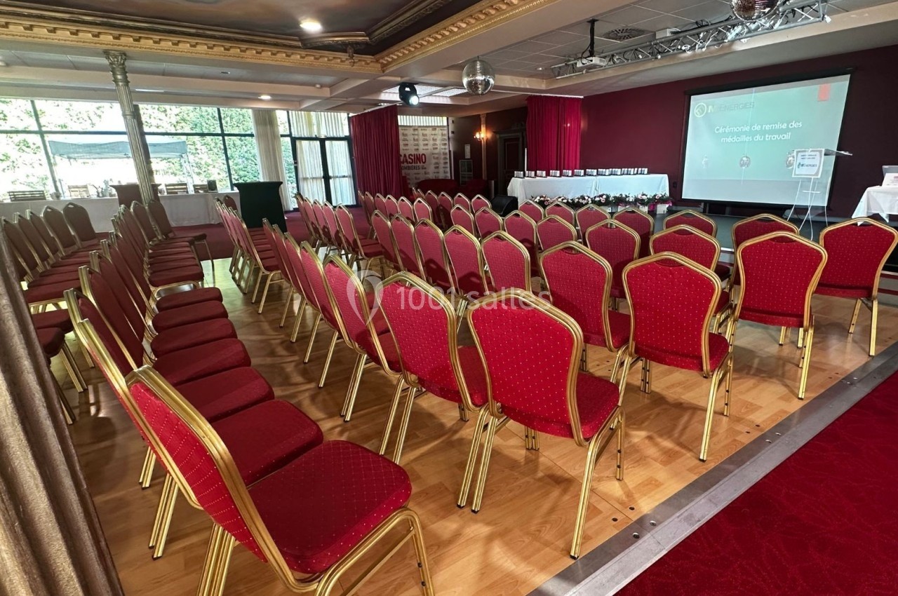 Salle de conférence avec des rangées de chaises rouges alignées face à un écran de projection et une scène.