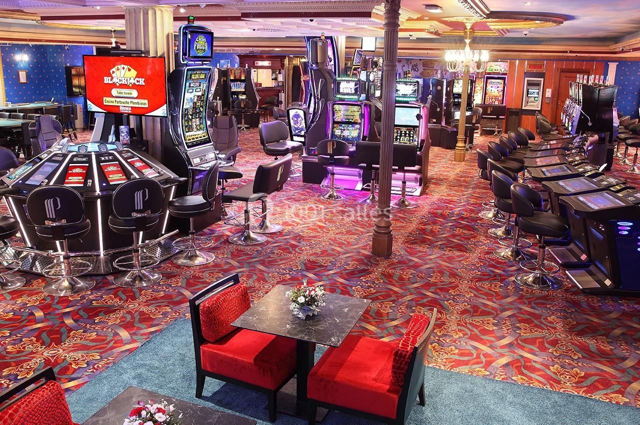 L'espace jeux avec le Black Jack et la Roulette Anglaise électroniques Salle de casino avec machines à sous, tables de jeux électroniques et sièges rouges sur un sol à motifs colorés.