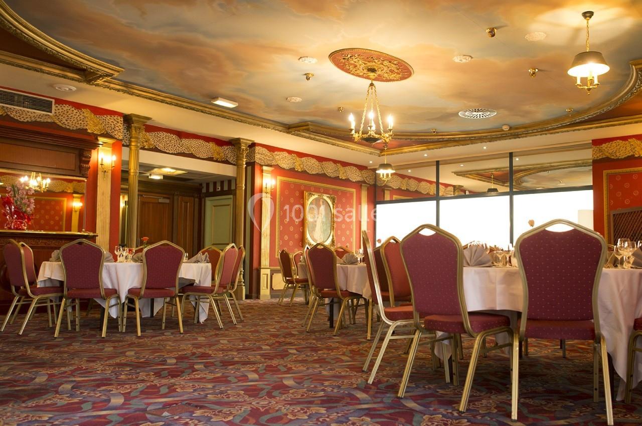 Le Salon de l'Empereur avant l'arrivée des convives Salle de réception élégante avec tables rondes dressées, chaises rouges et plafond décoré de fresques.