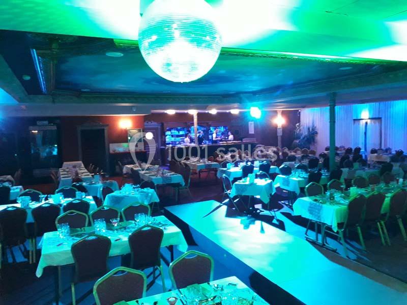 Les artistes prêts à entrer en piste Salle de réception éclairée avec des tables dressées, une piste centrale et un bar illuminé en arrière-plan.