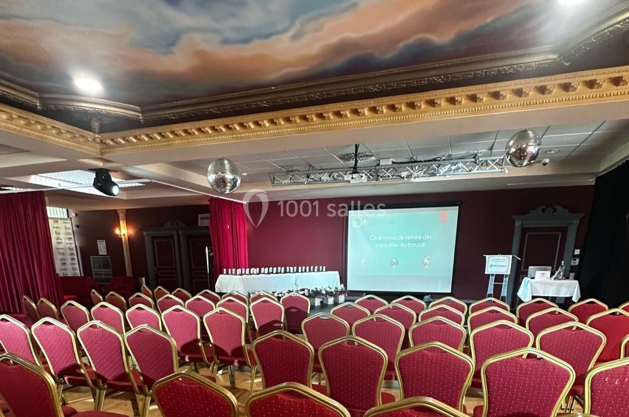 Salle de conférence avec rangées de chaises rouges, écran de projection allumé et décorations au plafond.