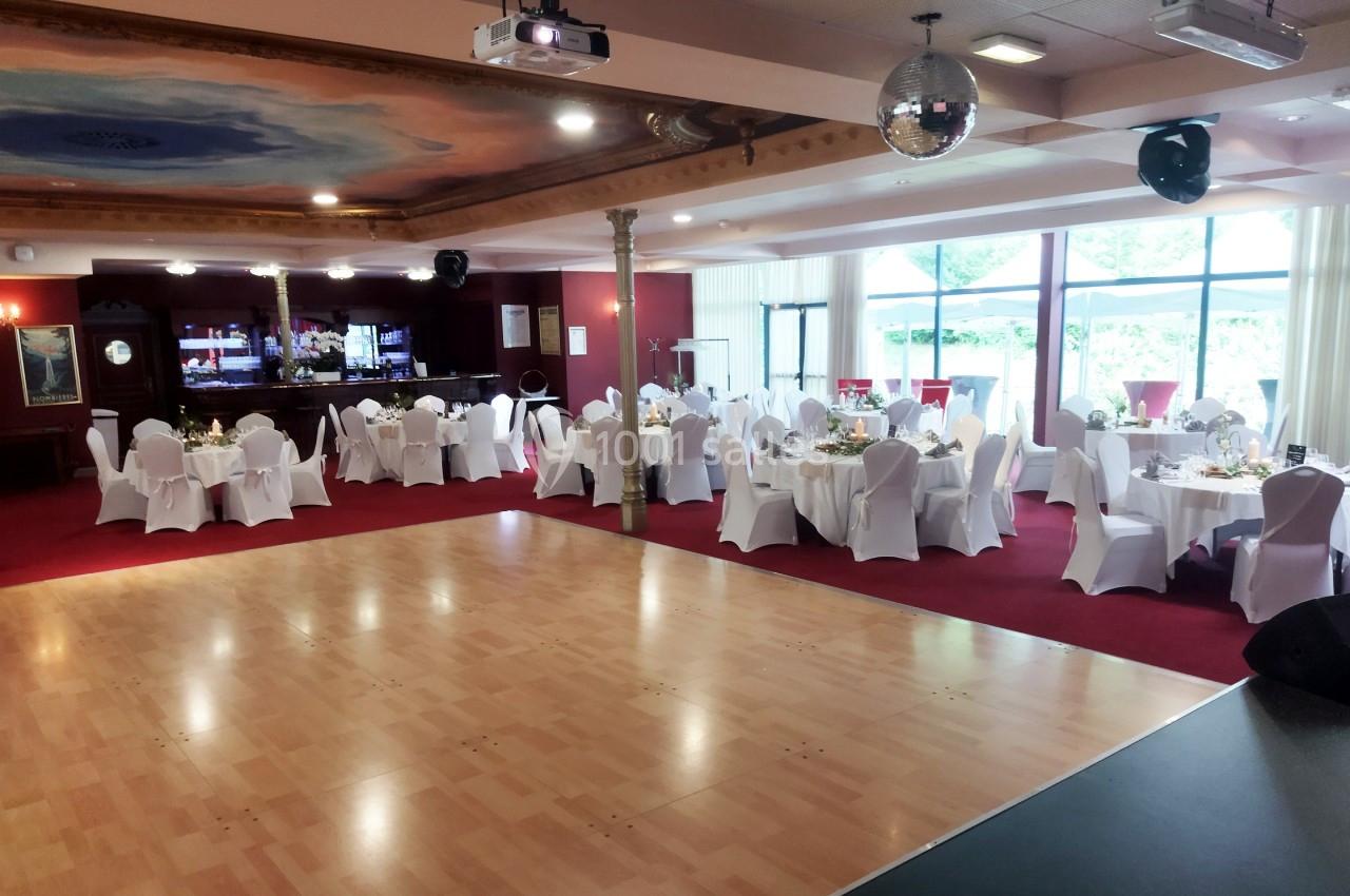 Repas et réception au Salon Impérial Salle de réception avec tables dressées, chaises blanches, piste de danse en bois et bar en arrière-plan.