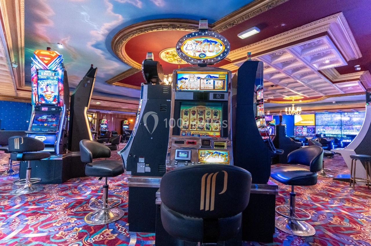 Salle des machines à sous du Casino Partouche de Plombières-les-Bains Salle de casino avec machines à sous illuminées, sièges rembourrés et plafond décoré de fresques.