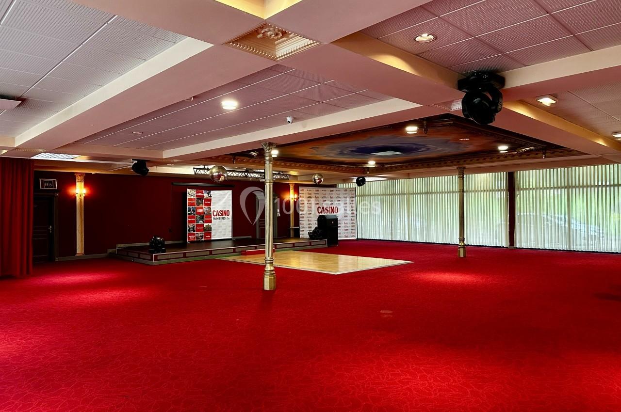 Salon Impérial aménageable selon l'évènement ou vide pour plus d'espace Salle spacieuse avec moquette rouge, scène surélevée, rideaux et grandes baies vitrées laissant entrer la lumière naturelle.