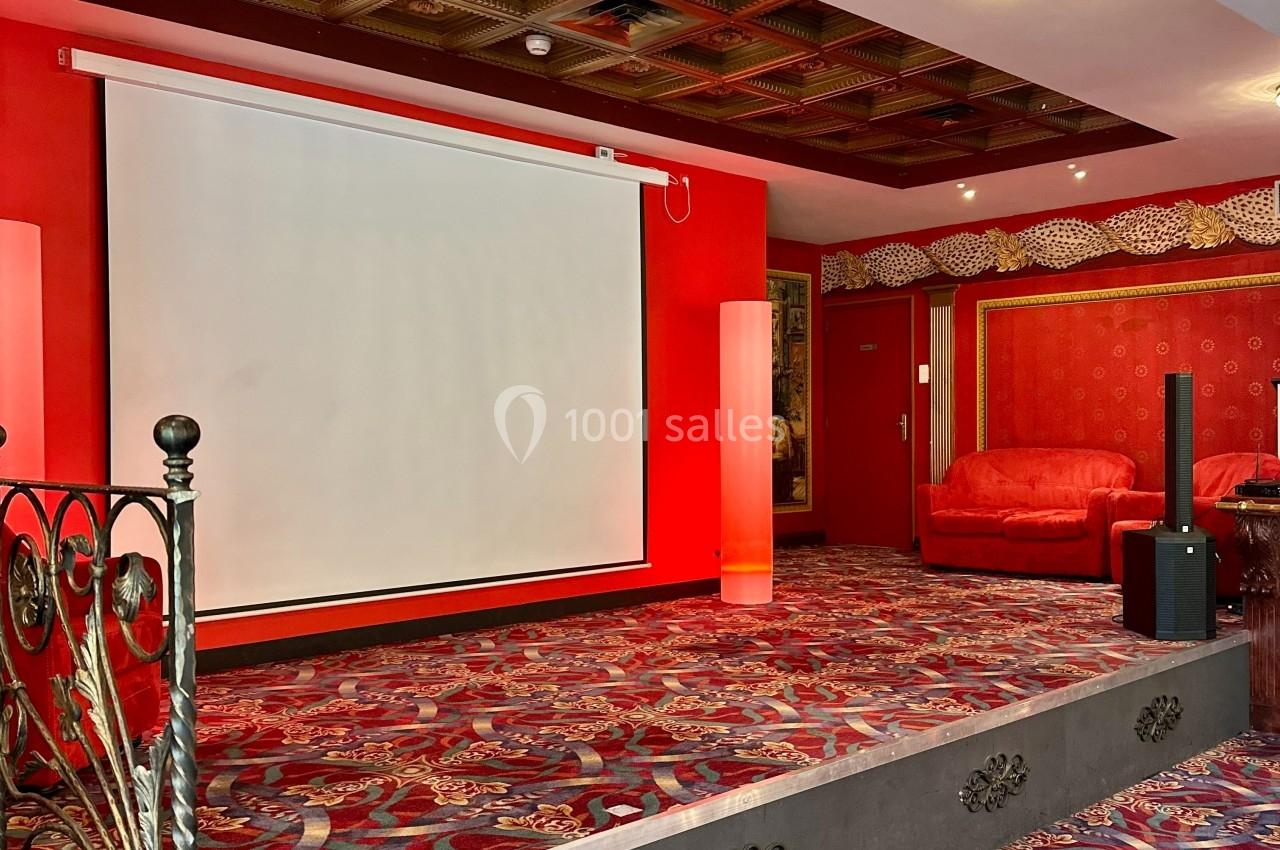Scène du Salon de l'Empereur Salle avec écran de projection, tapis rouge à motifs, fauteuils assortis et plafond à caissons décoratifs.