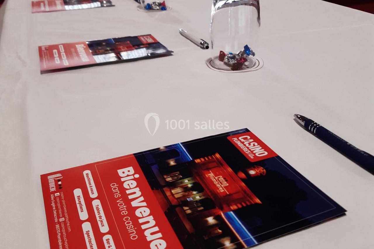 Tout est prêt pour la journée d'étude Brochures d'un casino posées sur une table blanche avec des stylos, un verre d'eau et des bonbons.