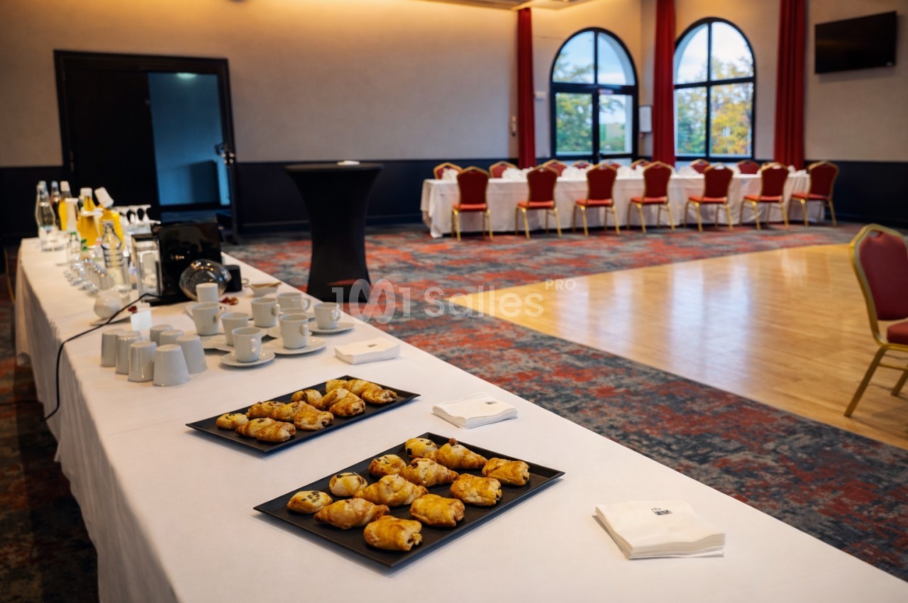 Table dressée avec viennoiseries, boissons et tasses dans une salle de réception lumineuse avec chaises alignées au fond.