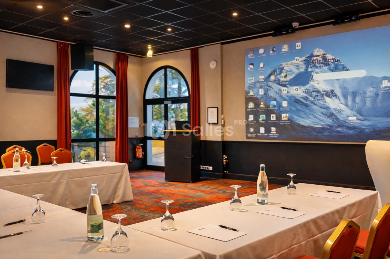 Salle de conférence équipée de tables, chaises, bouteilles d'eau, et écran de projection affichant un bureau d'ordinateur.