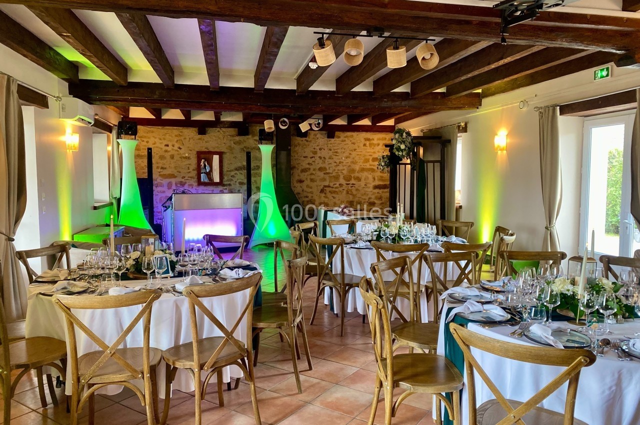 Salle de réception avec tables dressées, chaises en bois, éclairage coloré et murs en pierre apparente.