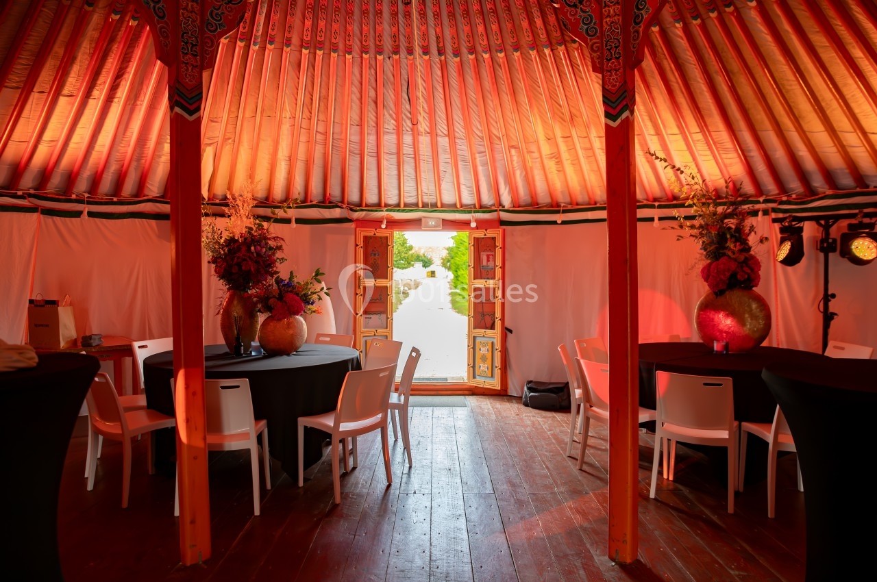 Intérieur d'une yourte décorée avec des tables, des chaises blanches et des arrangements floraux, éclairée par une lumière…