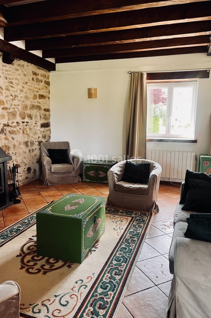 Salon rustique avec poutres apparentes, murs en pierre, cheminée, meubles verts décorés et fauteuils autour d'une table…