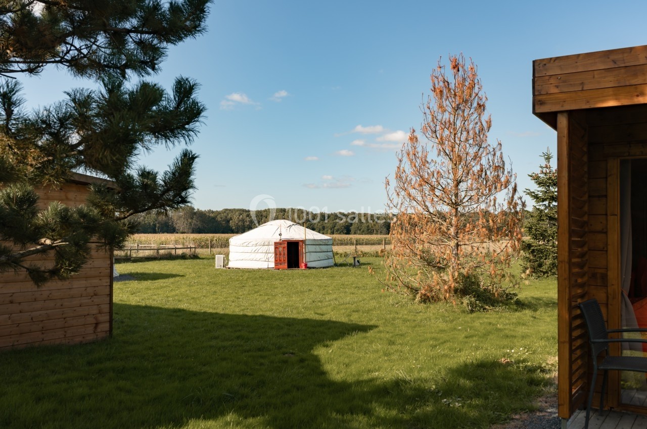 Yourte blanche au centre d'un jardin verdoyant, entourée d'arbres et de bâtiments en bois sous un ciel dégagé.