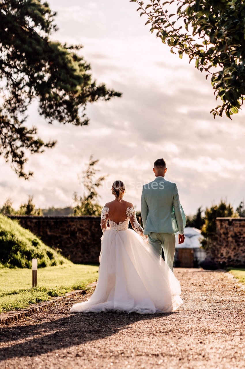 Un couple en tenue de mariage marche sur un chemin gravillonné, entouré de verdure et d'arbres sous un ciel nuageux.