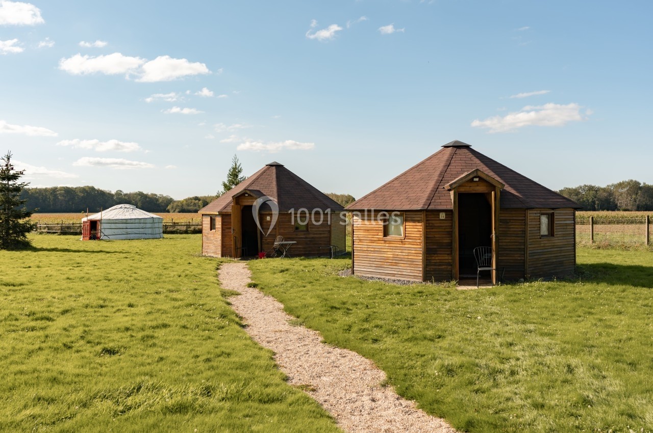 Chemin de gravier menant à deux cabanes en bois rondes dans un champ verdoyant sous un ciel dégagé.