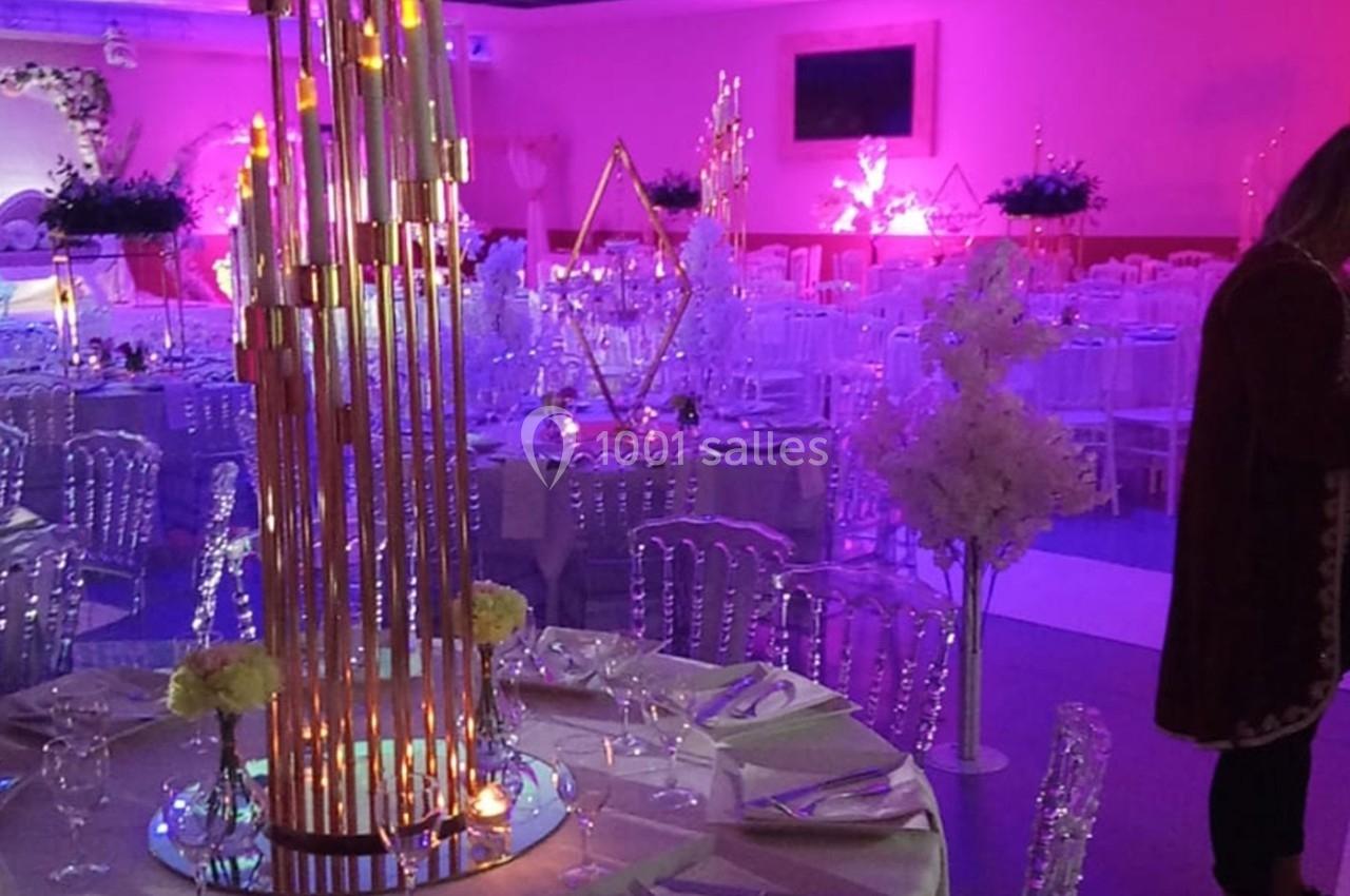Salle de réception décorée avec des tables dressées, des chaises transparentes et des éclairages roses et dorés.