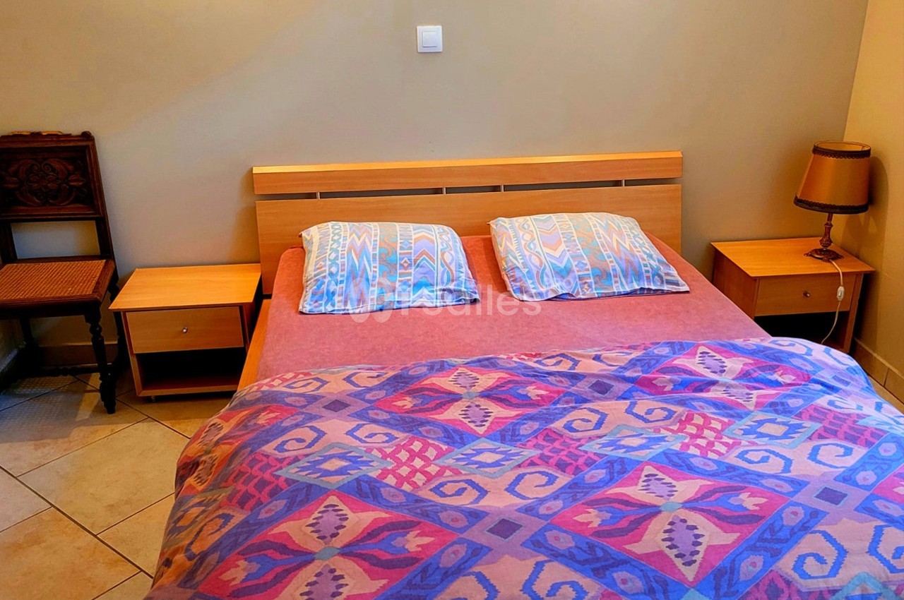Lit double avec une couverture colorée, deux oreillers, tables de chevet en bois et une lampe dans une chambre simple.