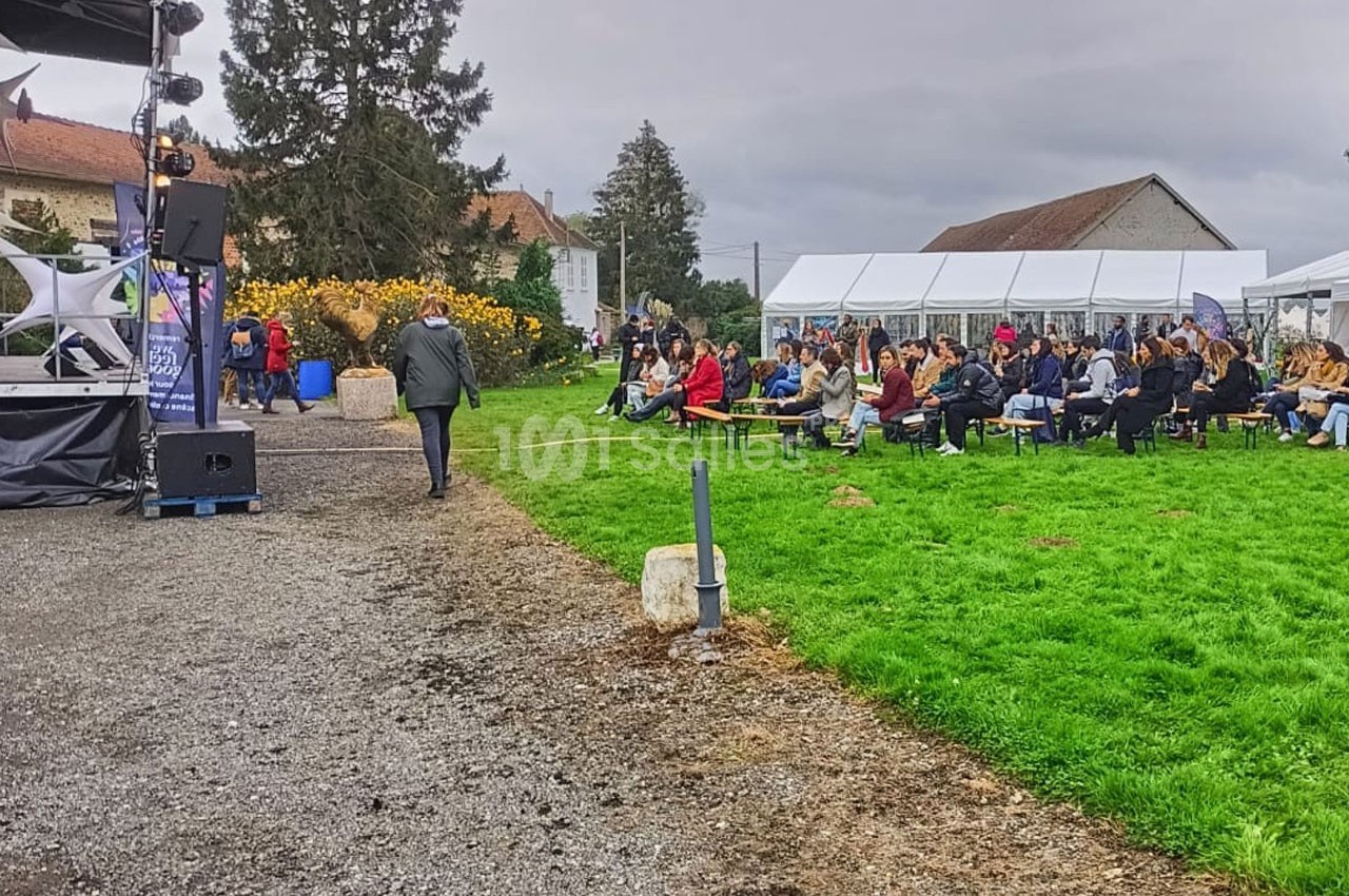 Un groupe de personnes assises en extérieur sous des chapiteaux, assistant à un événement près d'une scène.