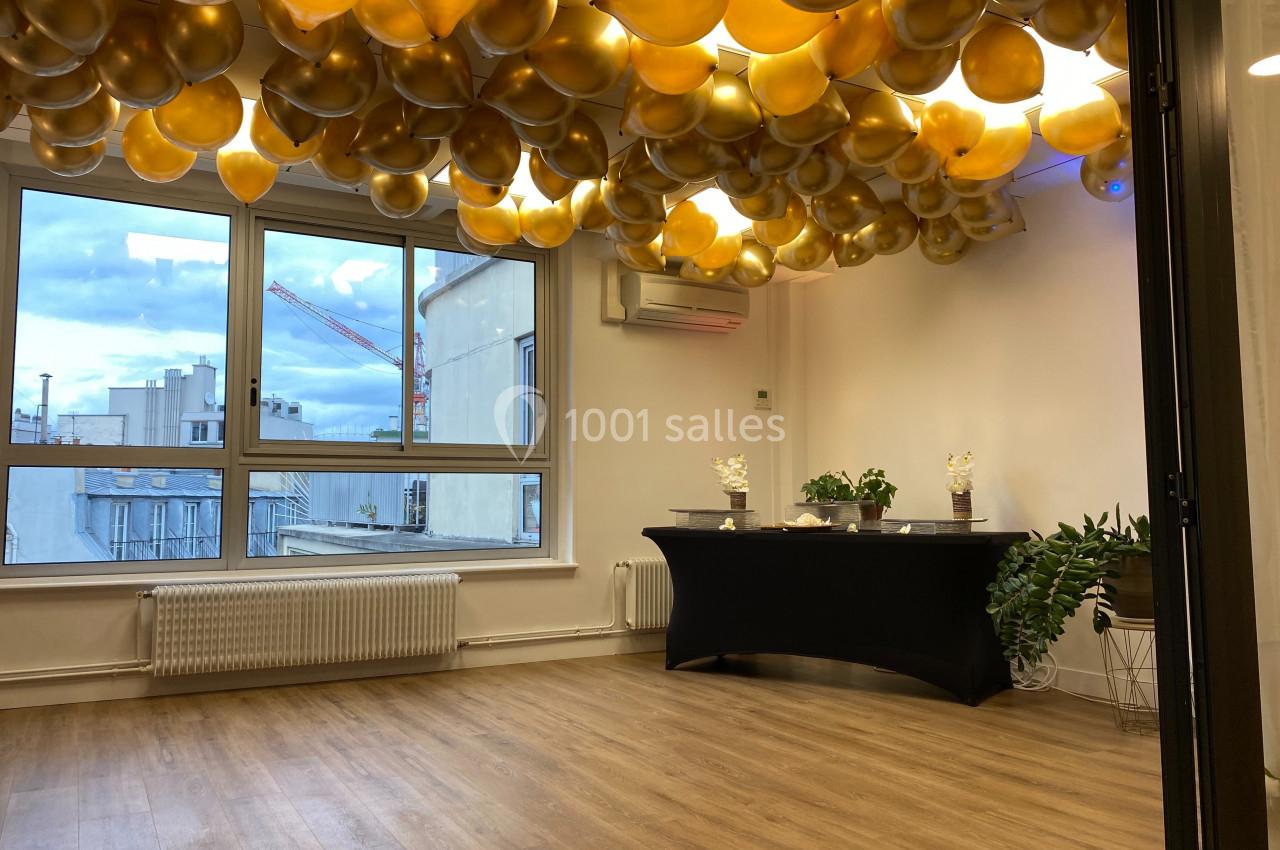 Salle lumineuse avec parquet, décorée de ballons dorés suspendus au plafond, vue sur des immeubles par de grandes fenêtres.