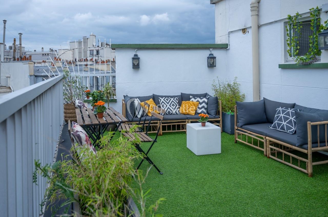 Terrasse aménagée avec mobilier en bois, coussins colorés, table, chaises et plantes sur un sol en gazon synthétique.