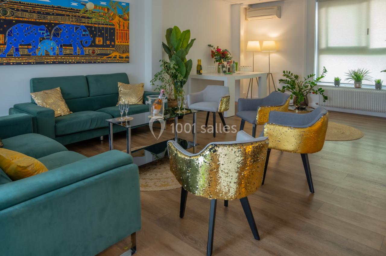 Salon moderne avec canapés verts, chaises dorées, plantes, tableau coloré et éclairage doux.