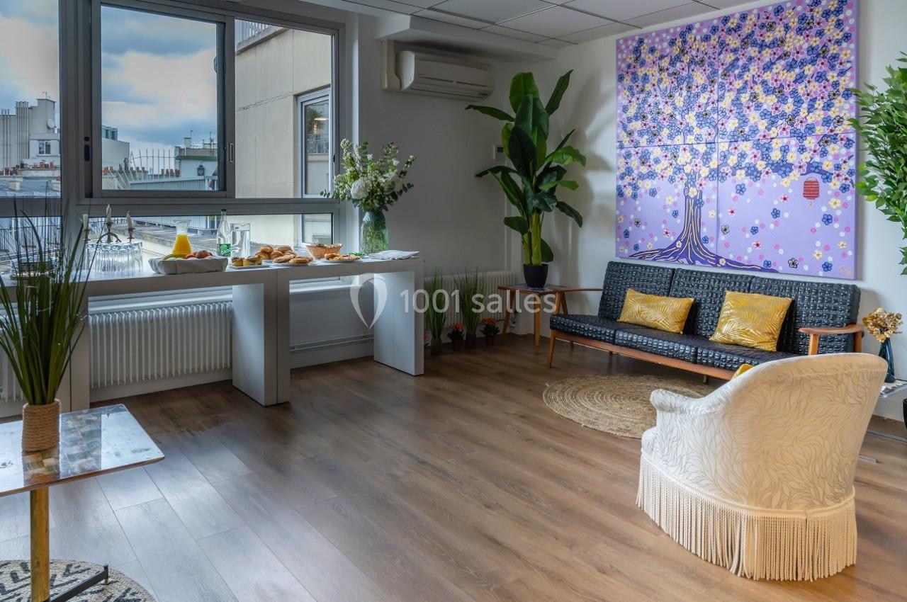 Salle lumineuse avec parquet, canapé, fauteuil, table dressée et plantes, donnant sur une vue urbaine par une grande fenêtre.