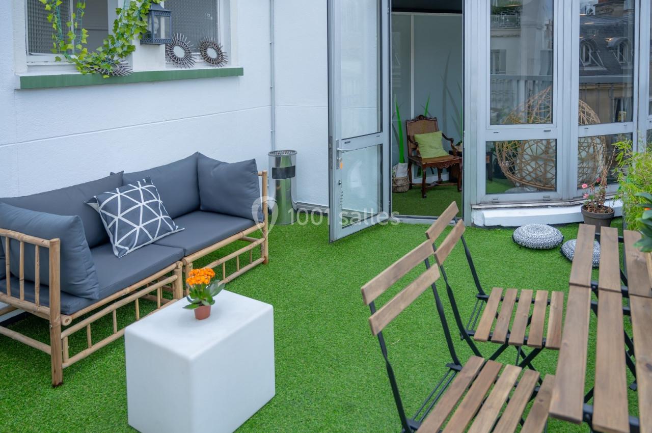 Terrasse avec gazon synthétique, canapé en bois, table et chaises pliantes, plantes et accès à une pièce intérieure.
