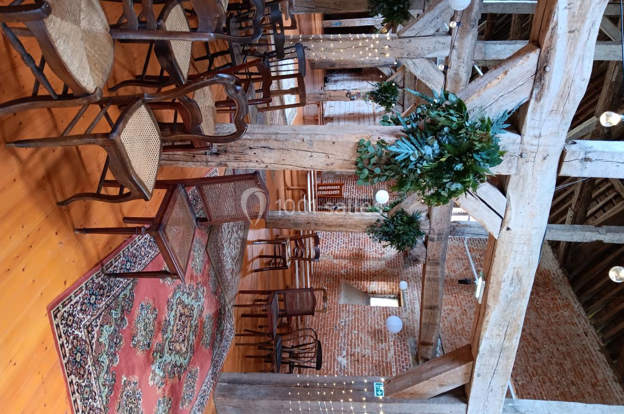 Salle rustique avec poutres apparentes, chaises en bois, tapis coloré et décorations végétales suspendues.